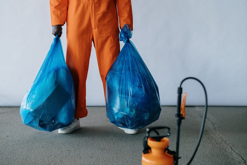 A person wearing a dark jacket and orange safety vest is pushing a blue trolley filled with recycling bins labeled for waste collection on a wet urban sidewalk, with trees and city buildings in the ba