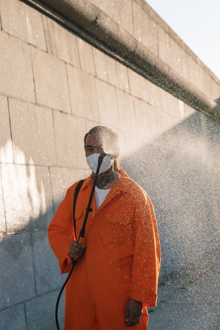 A person wearing a black hoodie, black pants, and white sneakers is engaged in surface cleaning by using a high-pressure washer to clean tiled wall-mounted panels in an industrial or commercial settin