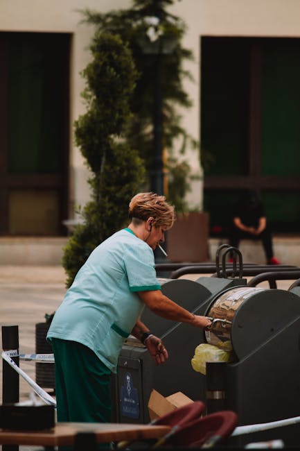 A street cleaner from Kingston Cleaner stands on a paved urban sidewalk during evening or early night, wearing a high-visibility orange uniform with reflective silver stripes for surface safety. The w