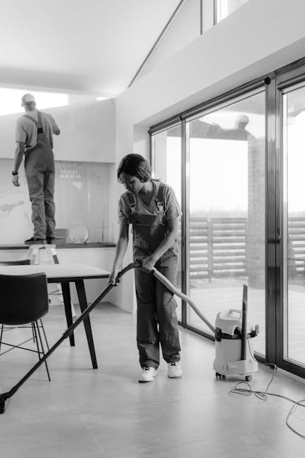 A professional cleaner from Kingston Cleaner is performing surface cleaning in a modern living space, using a vacuum cleaner and a hose attachment on a wooden floor. The room features large sliding glass doors leading to an outdoor balcony, allowing natural light to illuminate the area. A dining table with chairs is visible, and a second cleaner is in the background on a step ladder, attending to high surfaces on the cabinet or wall. The surfaces appear well-maintained and dust-free, with the cleaner focusing on thorough sanitisation. The setting reflects a clean, organized environment suited for deep cleaning and domestic hygiene, demonstrating the company's expertise in end-of-tenancy cleaning services using appropriate materials and tools for a comprehensive clean.