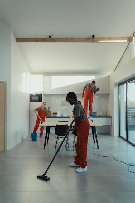 A team of three professional cleaners from Kingston Cleaner are performing surface cleaning and deep cleaning in a modern, spacious kitchen area. One cleaner in the foreground is using a vacuum cleaner on the tiled floor, which has a smooth, light grey ceramic surface, ensuring thorough dust removal. In the background, another cleaner is wiping the countertop, which appears to be made of light-colored stone or composite material, with a cloth or spray bottle, emphasizing attention to hygiene and sanitisation. The third cleaner is standing on a step ladder near the wall, cleaning high surfaces or upper cabinets, with a spray bottle in hand. The room is illuminated by natural sunlight coming through large glass sliding doors, casting bright, even light across the clean, minimalist space. The ceiling is painted light grey, with a wooden beam for architectural detail, and the walls are white, contributing to a fresh and hygienic appearance. The scene reflects professional domestic cleaning practices aimed at maintaining high standards of cleanliness in a residential setting, as provided by Kingston Cleaner, aligning with services such as end of tenancy cleaning and same-day quotes and rates.