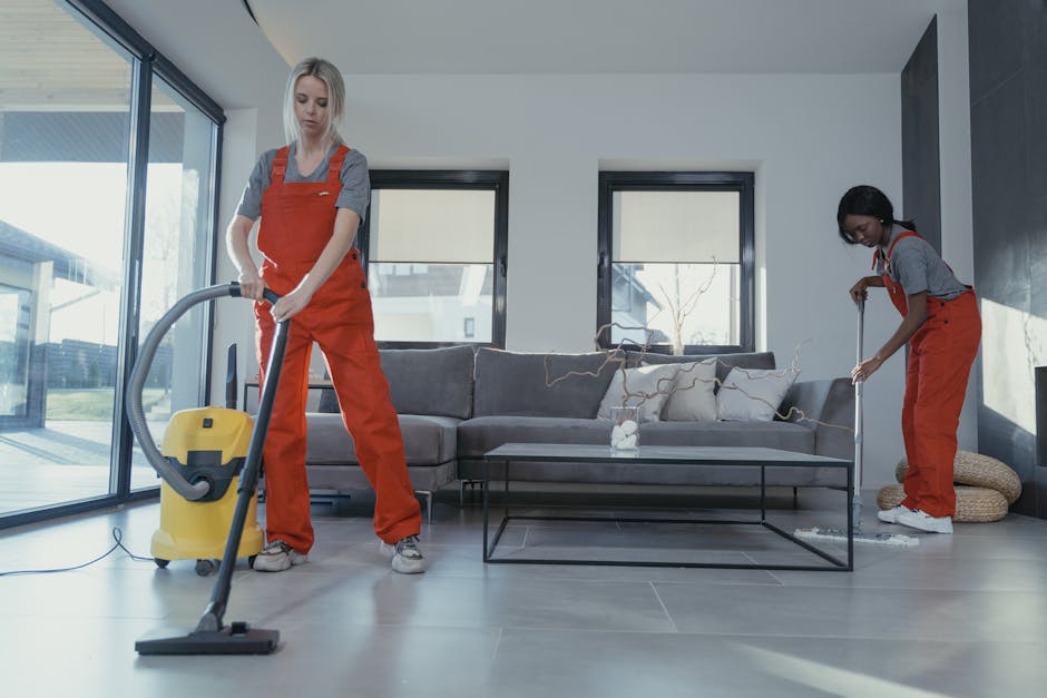 A professional cleaner from Kingston Cleaner is performing surface cleaning in a modern living space, using a vacuum cleaner and a hose attachment on a wooden floor. The room features large sliding glass doors leading to an outdoor balcony, allowing natural light to illuminate the area. A dining table with chairs is visible, and a second cleaner is in the background on a step ladder, attending to high surfaces on the cabinet or wall. The surfaces appear well-maintained and dust-free, with the cleaner focusing on thorough sanitisation. The setting reflects a clean, organized environment suited for deep cleaning and domestic hygiene, demonstrating the company's expertise in end-of-tenancy cleaning services using appropriate materials and tools for a comprehensive clean.