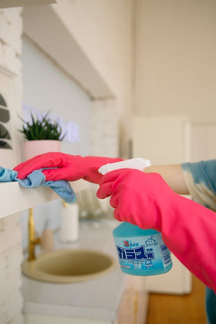 A woman wearing a black cleaning uniform is using a yellow-handled duster to clear dust from the edge of a large flat-screen television mounted on a plain white wall in a residential setting. The tele